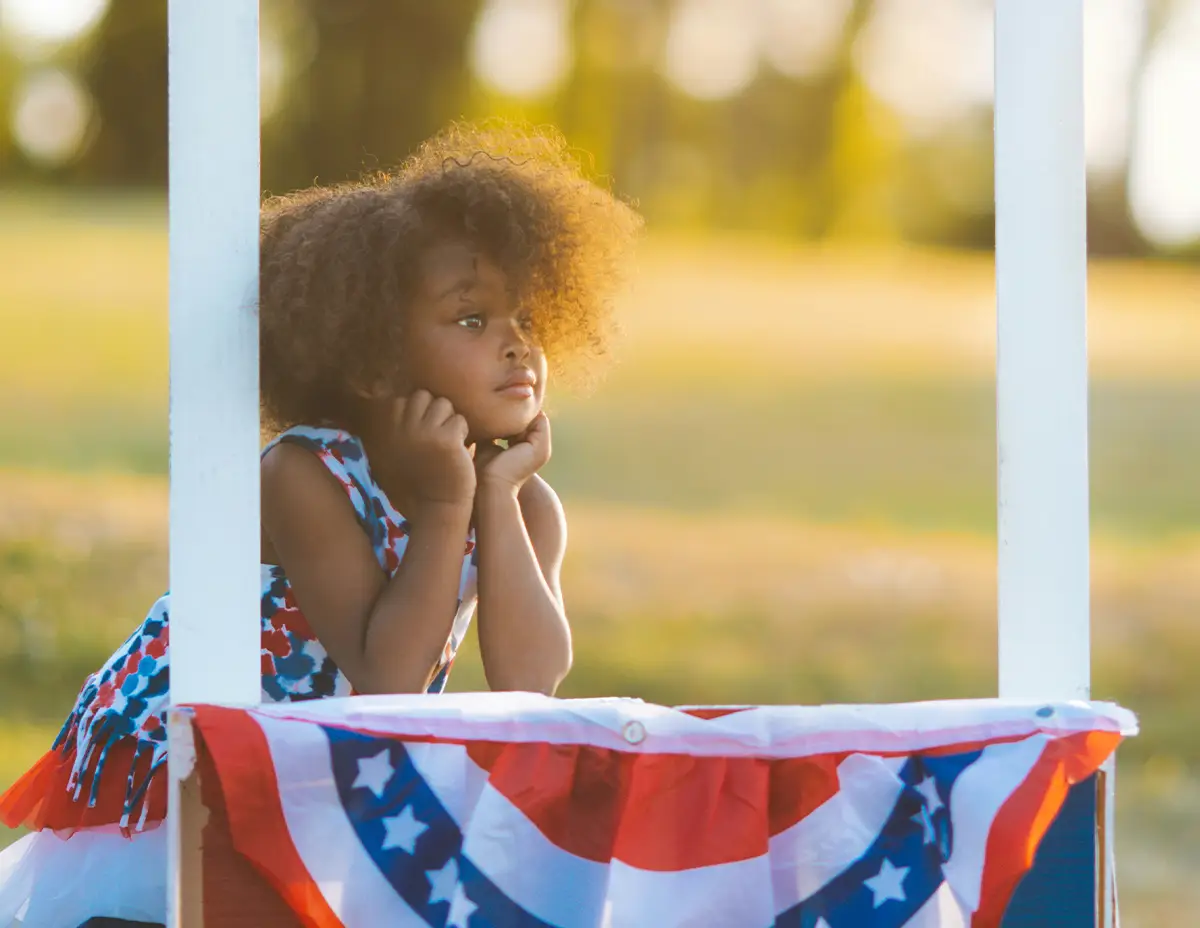 Young child staring toward sunset leaning over red, white and blue tablecloth.