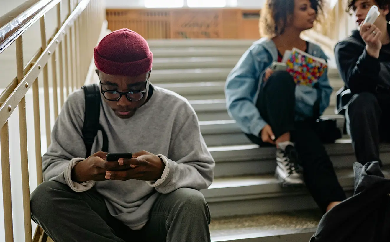 High school student sitting away from friends, isolated with face buried in phone screen.