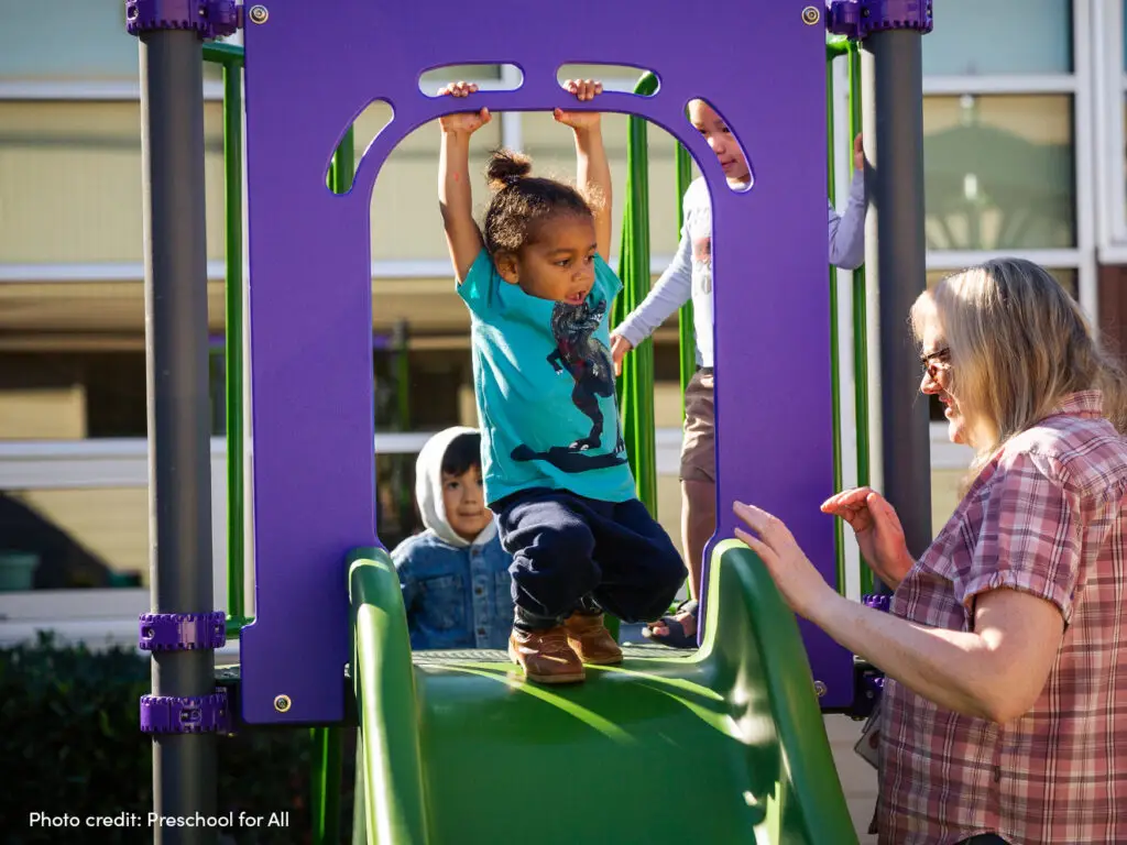 Young child going down slide and an adult approaching to help them.