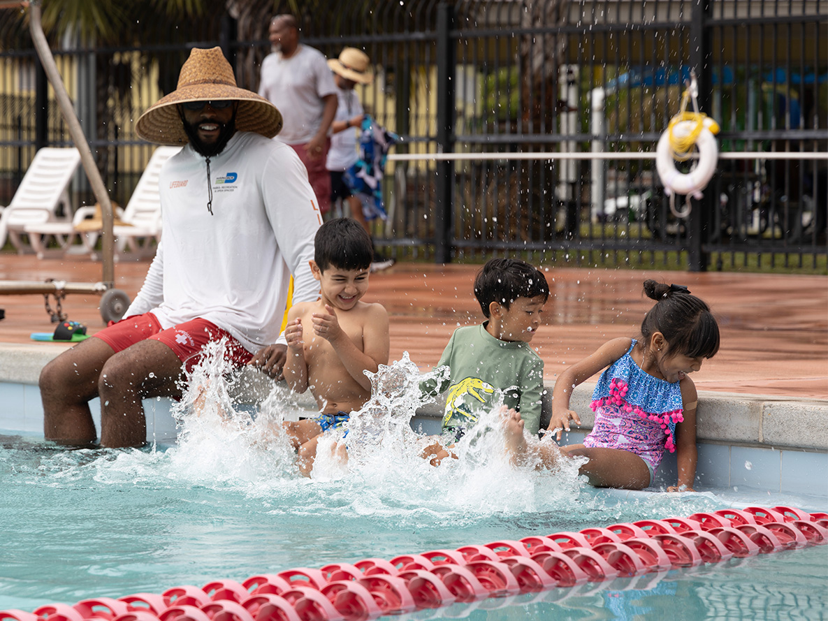 Adult sitting with several kids kicking pool water and splashing each other